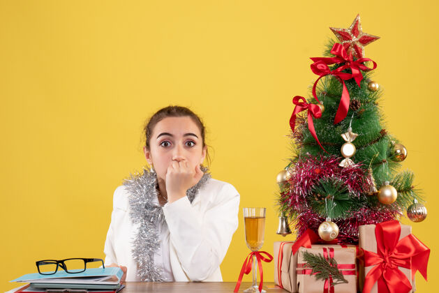 背景正面图：女医生坐在黄色背景的桌子后面 还有圣诞树和礼品盒快乐情绪微笑