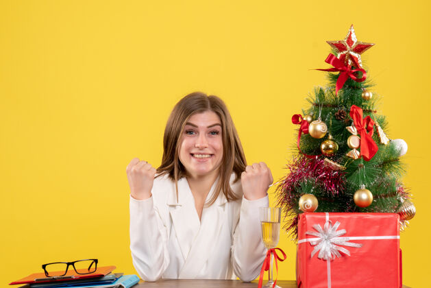 人正面图：女医生坐在黄色背景的桌子前 放着圣诞树和礼品盒生活成人职业