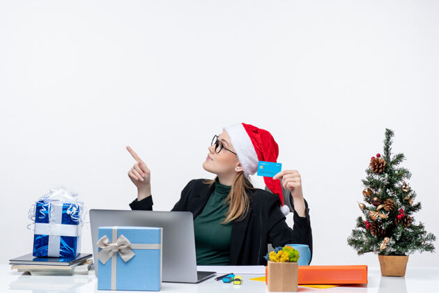 坐着一个戴着圣诞老人帽子戴着眼镜的漂亮女人坐在桌旁 手里拿着圣诞礼物和银行卡在办公室的镜头里迷人帽子女人