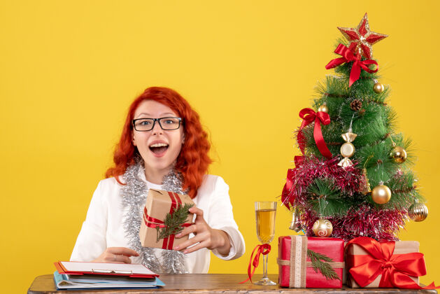 坐着前视图：女医生坐在桌子后面 黄色背景上有圣诞礼物背景医生生活