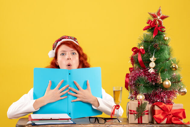 坐着正面图：女医生坐在桌子后面 手里拿着文件 背景是黄色的圣诞树和礼盒女医生快乐女性