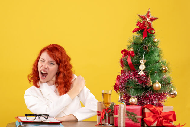 背景正面图：女医生坐在桌子后面 拿着圣诞礼物 黄色背景上的树礼物快乐安排