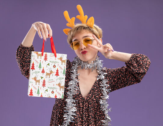 头带自信的年轻漂亮女孩戴着驯鹿鹿角头带 脖子上戴着金箔花环 戴着眼镜 拿着圣诞礼品袋 看着相机 在紫色背景上做着和平的手势抱着快乐包
