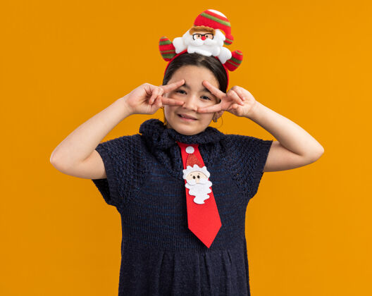 圣诞节快乐的小女孩穿着针织连衣裙 头上系着一条红色领带 戴着一个有趣的圣诞戒指 看着摄像机 在橙色的背景下 眼睛附近有一个v型标志眼睛近小
