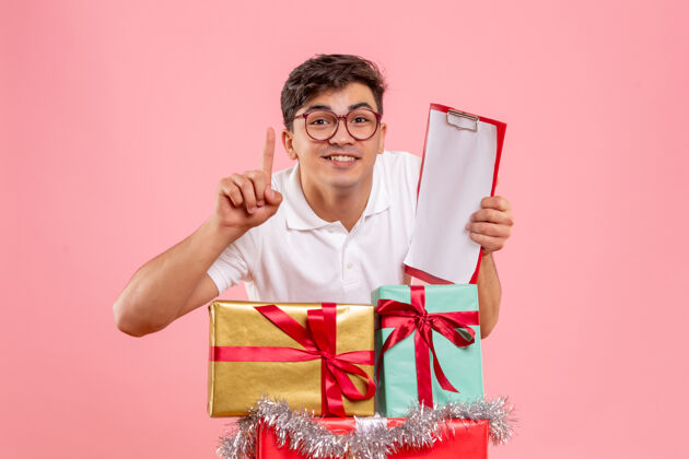礼物年轻人圣诞节礼物的正面图拿着文件纸条微笑着贴在粉红色的墙上前面举行节日