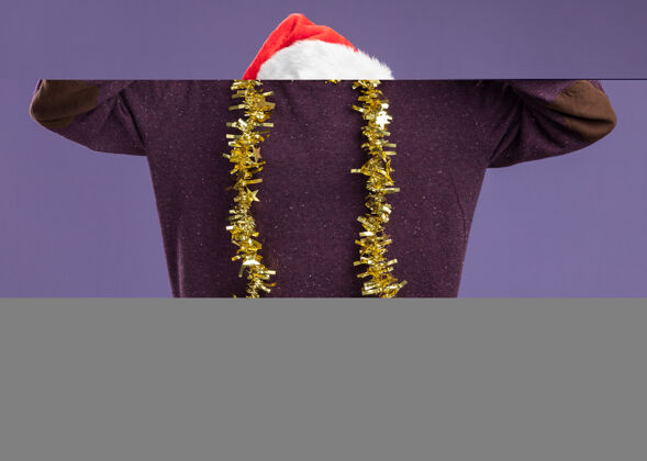 圣诞老人年轻的留着胡子的男子戴着圣诞老人帽 脖子上戴着金属丝 看着摄像机 他愉快地微笑着 紫色背景上立着v字标志圣诞快乐年轻脖子