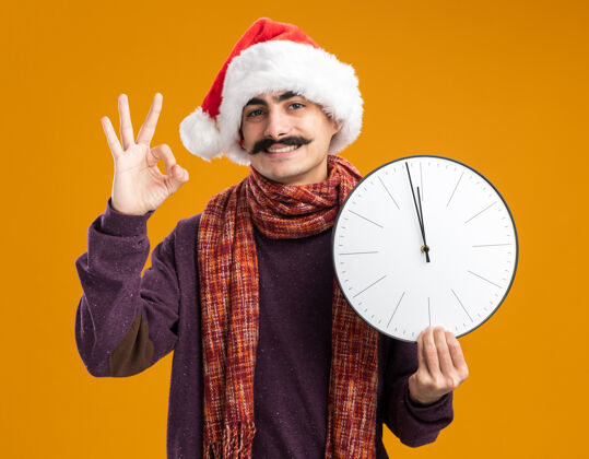 围着快乐的留着胡子的男人戴着圣诞老人帽 脖子上围着暖和的围巾 手里拿着时钟 微笑着在橙色的背景上立着ok标志展示脖子抱着