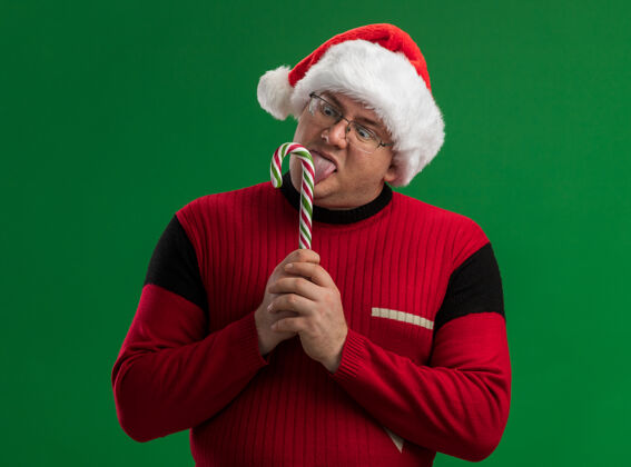 帽子印象深刻的成年男子戴着眼镜和圣诞老人的帽子 拿着圣诞糖果手杖舔着它 孤立在绿色的背景上甜甜的手杖舔着