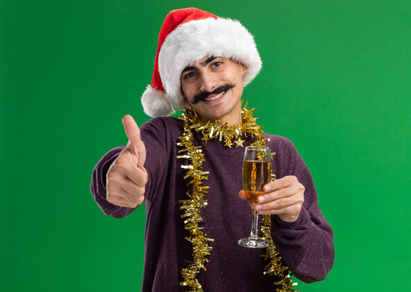 高兴年轻的留着胡子的男人戴着圣诞老人帽子 脖子上戴着金属丝 手里拿着一杯香槟 看着摄像机 兴高采烈地微笑着 竖起大拇指站在绿色的背景上玻璃杯圣诞节帽子
