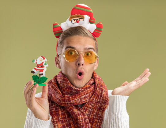 圣诞老人印象深刻的年轻帅哥戴着圣诞老人的头带和围巾拿着圣诞老人玩具显示空手孤立在橄榄绿的墙上穿着圣诞老人围巾