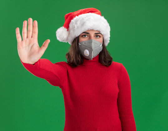 手势穿着红色毛衣 戴着圣诞老人帽的小女孩戴着护面面具 严肃地做着停下来的手势 手站在绿色的墙上认真圣诞老人制作