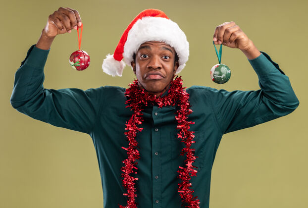 站着一个戴着圣诞帽 戴着花环 拿着圣诞球的非裔美国人站在绿色的墙上 既惊讶又困惑帽子花环惊讶