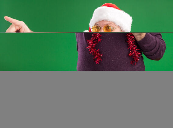 日期令人印象深刻的中年男子戴着圣诞帽 脖子上戴着金箔花环 戴着眼镜 手里拿着圣诞树玩具 看着日期 指着绿色墙上孤立的一面侧面周围印象深刻