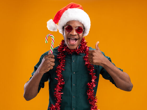 站立戴着圣诞帽戴着花环戴着眼镜拿着糖果手杖微笑着竖起大拇指站在橙色的墙上的非裔美国人圣诞老人非洲花环