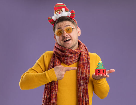 立方体快乐的年轻人穿着黄色高领毛衣 戴着暖和的围巾和眼镜 戴着滑稽的帽沿 头上戴着圣诞老人 在紫色的墙上展示着玩具立方体和新年的日期高领毛衣边缘围巾