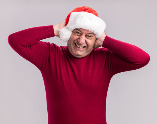 表情中年男子戴着圣诞老人的帽子 站在白色的背景上 双手合上耳朵 表情烦躁圣诞老人圣诞节帽子