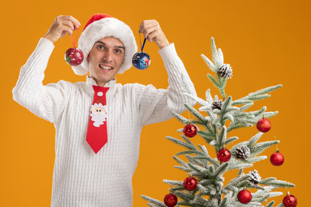 帅气快乐的年轻帅哥戴着圣诞帽 打着圣诞老人的领带 站在装饰好的圣诞树旁 手里拿着圣诞球饰物 脑袋附近被隔离在橙色的墙上球圣诞节装饰品