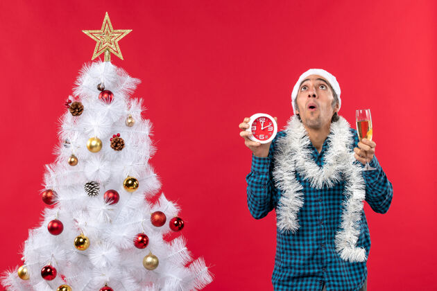 时钟一个戴着圣诞老人帽子 拿着一杯葡萄酒和圣诞树旁的钟的关心的年轻人小伙子葡萄酒圣诞