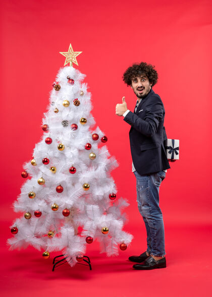 圣诞节快乐的年轻人拿着他的礼物站在红色的右边装饰的白色圣诞树旁做着ok的手势站着庆祝手势