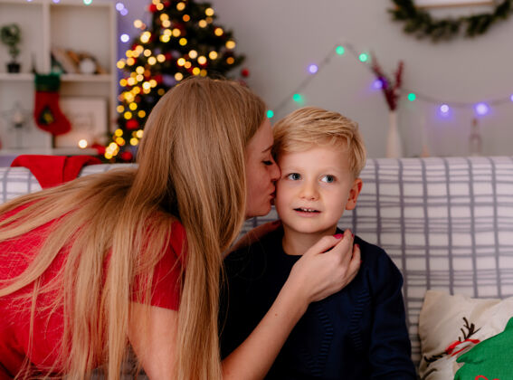 接吻在一间装饰华丽的房间里 身着红色连衣裙的快乐妈妈亲吻坐在沙发上的孩子 背景是圣诞树衣服快乐坐