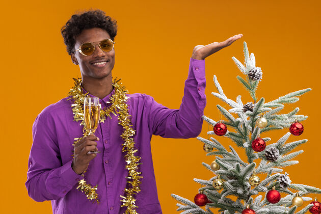 手微笑着的年轻的非裔美国人戴着一副戴着金箔花环的眼镜 站在装饰好的圣诞树旁 手里拿着一杯香槟圣诞树戴着新年