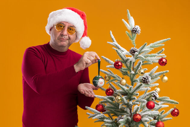 红色中年男子戴着圣诞老人帽 深红色高领毛衣 戴着黄色眼镜 带着怀疑的微笑装饰圣诞树深色黄色年龄