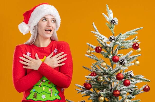 金发穿着圣诞毛衣 戴着圣诞帽的金发女郎站在橙色背景下的圣诞树旁 双手放在胸前 神采奕奕地看着一边圣诞老人抱着毛衣