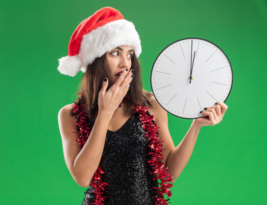 时钟年轻漂亮的女孩戴着圣诞帽 脖子上戴着花环 手被隔离在绿色的墙上 手里拿着挂钟 眼睛盯着挂钟盖害怕花环