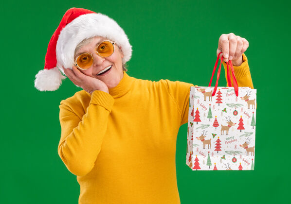 老人戴着太阳眼镜 戴着圣诞老人帽子的兴奋的老妇人把手放在脸上 手里拿着一个纸礼包 在绿色的背景上隔离开来 还有复印空间圣诞老人帽子脸