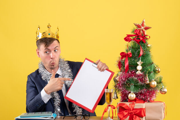 指向前视图的商人指着剪贴板坐在桌旁圣诞树和黄色的礼物坐着办公室树