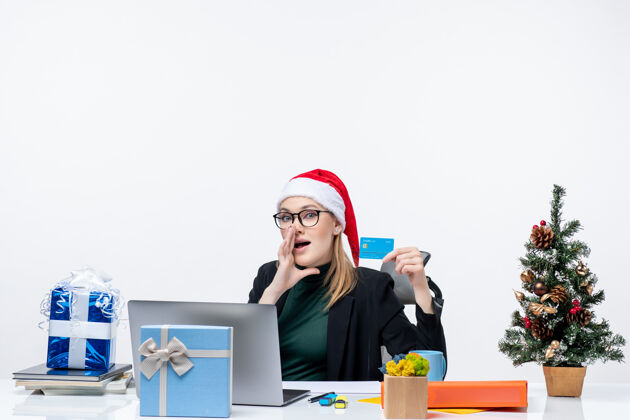 女商人戴着圣诞老人帽戴着眼镜的女商人坐在桌子旁 手里拿着银行卡 给办公室里的人打电话企业家礼物树
