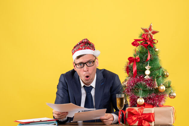 黄色快乐的商人坐在圣诞树旁的桌子旁查看文件和黄色的礼物桌子礼物男人