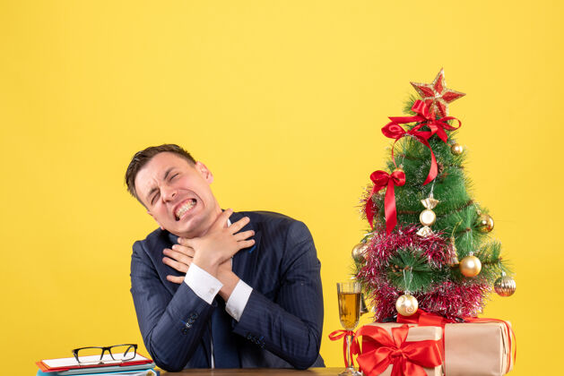 礼物愤怒的男人双手勒死自己坐在圣诞树旁的桌子前 黄色的墙上挂着礼物桌子人商人
