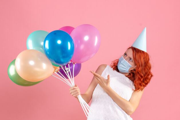 气球前视图中的年轻女子拿着五颜六色的气球在无菌面具上粉红色的墙壁乐趣Covid前面