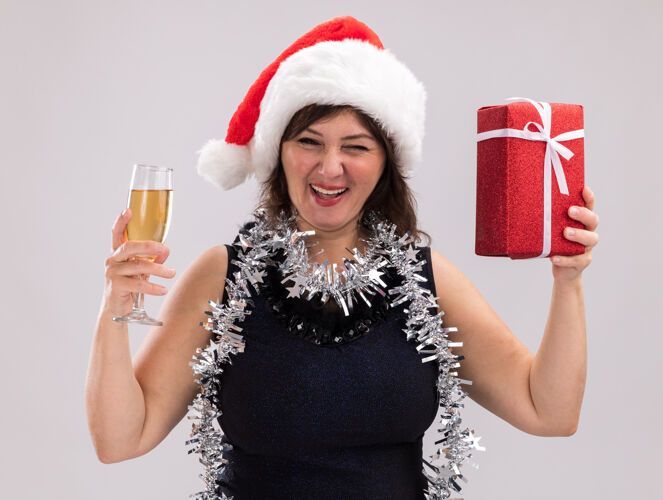 礼物快乐的中年妇女戴着圣诞帽 脖子上戴着金箔花环 手里拿着圣诞礼包 手里拿着一杯香槟 看着相机在白色背景上眨眼包装中年帽子