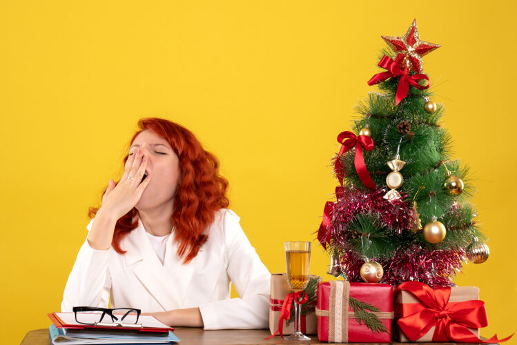 树女工人坐在桌子后面 手里拿着圣诞礼物 树在黄色上打着哈欠女工人工人圣诞节