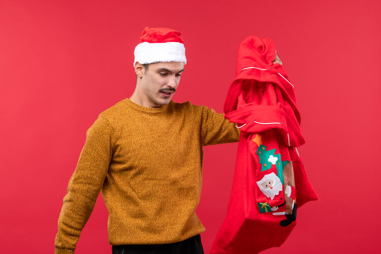 帽子红墙上拿着礼物包的年轻人的正视图圣诞节男性男人