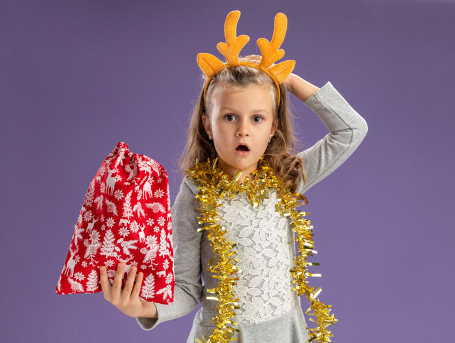 包困惑的小女孩戴着圣诞发箍 脖子上戴着花环 手放在头上 手放在蓝色背景上圣诞头花环