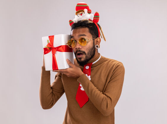 非洲一个非洲裔美国人 穿着棕色毛衣 头戴圣诞老人圈 打着有趣的红色领带 手里拿着礼物 站在白色背景下惊讶地看着相机头站着惊讶