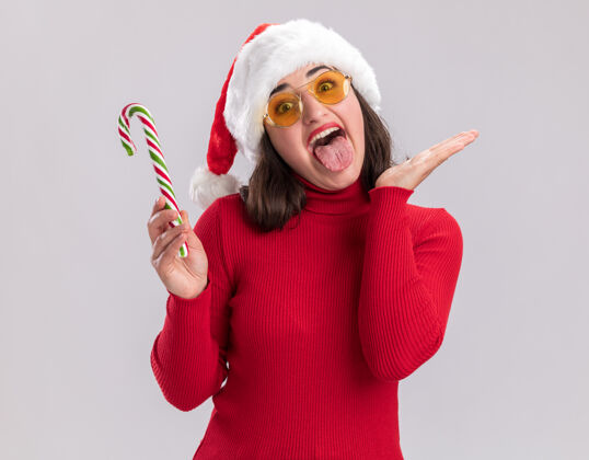 女孩穿着红毛衣 戴着圣诞帽 戴着眼镜 手里拿着糖果拐杖的年轻女孩 快乐而羞涩地伸出舌头站在白色的背景上年轻站着手杖