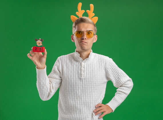 养可疑的年轻帅哥戴着驯鹿鹿角头带戴着眼镜拿着雨鹿鹿角玩具手放在腰上隔离在绿色的墙上圣诞节年轻鹿角