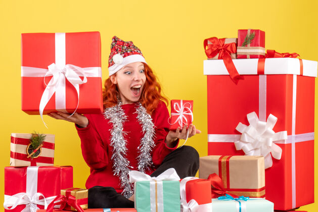 快乐坐在黄色墙上的圣诞礼物周围的年轻女子的正视图情绪前面盒子
