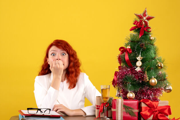 坐着女工人坐在桌子后面 手里拿着圣诞礼物 圣诞树紧张地挂在黄色的树上人礼物主任