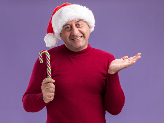 站着快乐的中年男人戴着圣诞老人的帽子拿着糖果手杖看着相机 脸上带着微笑 举起手臂站在紫色的背景上手杖胳膊帽子