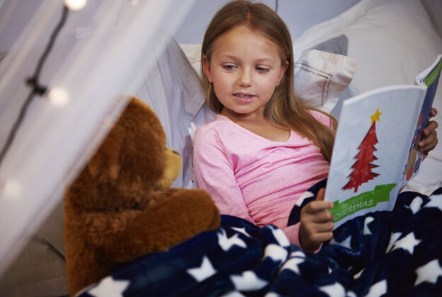 玩和泰迪熊一起读故事书的女孩抱着圣诞节晚上熊