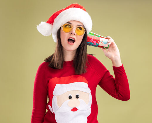 圣诞老人好奇的年轻漂亮的白人女孩 穿着圣诞老人的毛衣 戴着圣诞帽 戴着眼镜 手里拿着塑料圣诞杯 耳边听着谈话杯子举行年