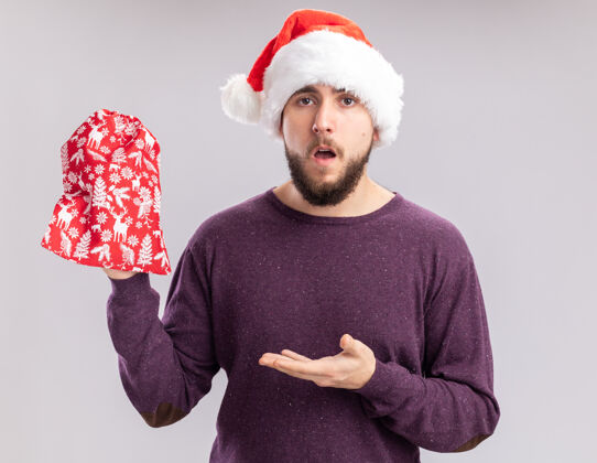站着穿着紫色毛衣 戴着圣诞帽的年轻人手里拿着红包 手里拿着礼物 站在白色的背景上 手臂看起来很困惑帽子包礼物