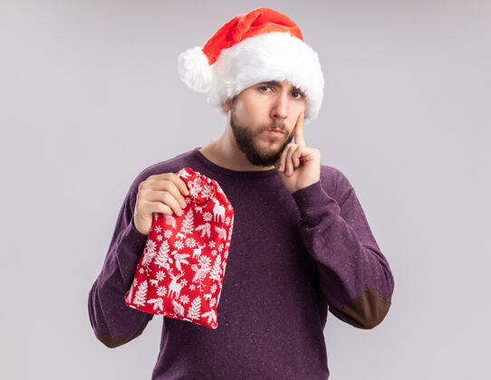 站着穿着紫色毛衣 戴着圣诞帽的年轻人手里拿着红包 手里拿着礼物 站在白色背景下 带着怀疑的表情看着镜头圣诞老人毛衣帽子