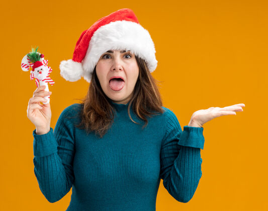 帽子恼怒的成年白人妇女戴着圣诞帽 把舌头伸出来 拿着糖果手杖孤立地放在橙色背景上 留着复制空间圣诞老人成人持有