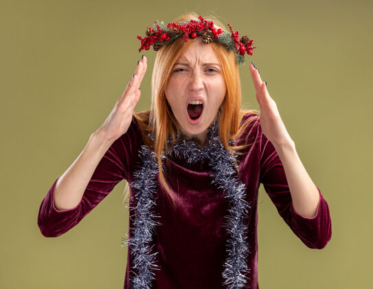 圣诞节愤怒的年轻漂亮女孩穿着红裙子 脖子上戴着花环和花环 隔离在橄榄绿的墙上礼服脖子花环